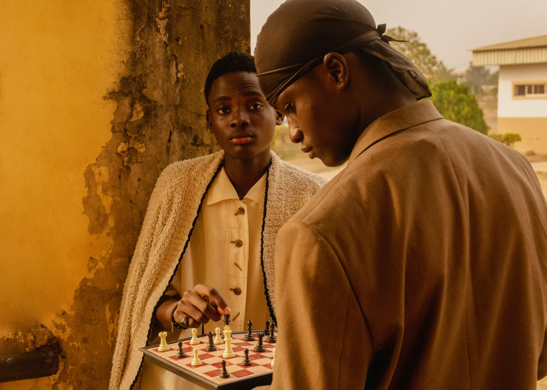 Two young adults engage in a chess game outdoors in Ado Ekiti, Nigeria, captured in warm tones.